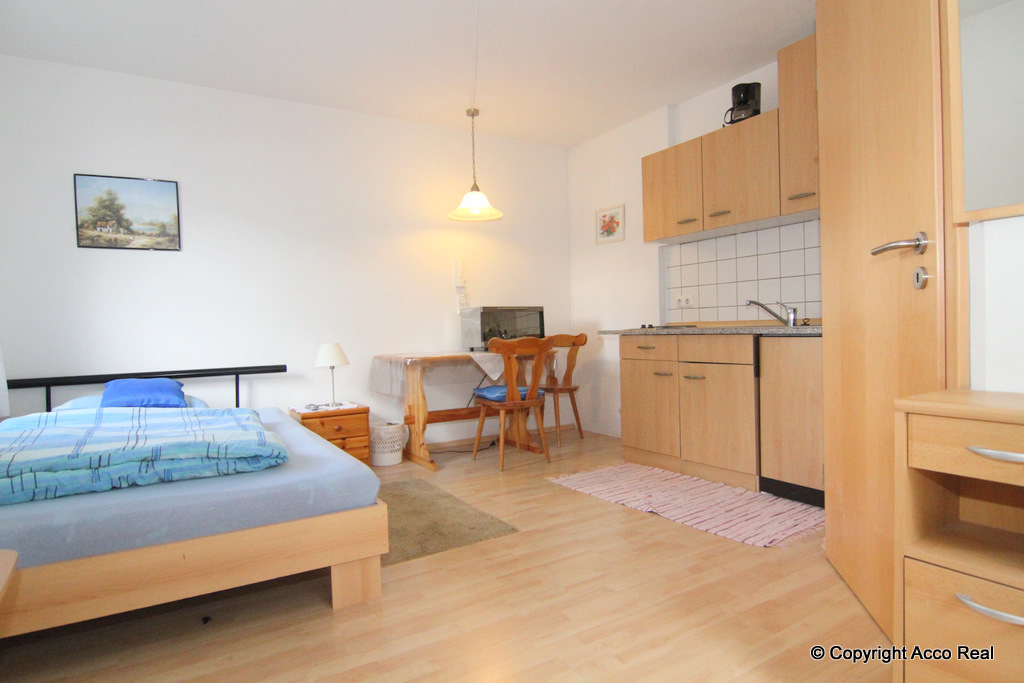 Wohn- und Schlafraum mit Bett, Esstisch, Holzstühlen und Küchenzeile mit Holzfronten.