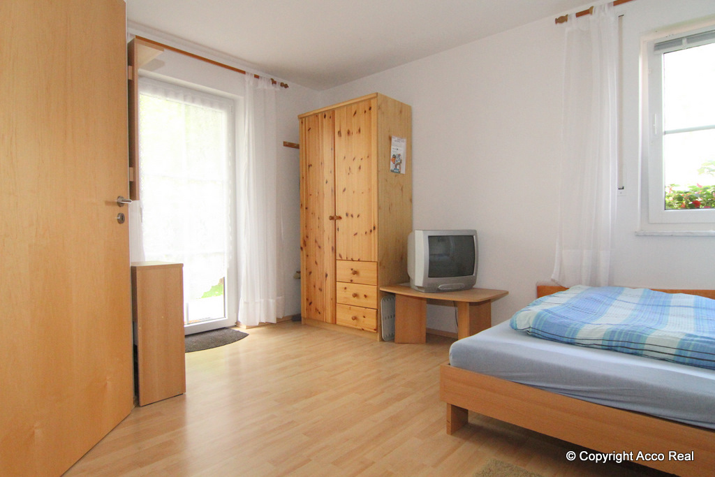 Möbliertes Zimmer mit Bett, Holzschrank, Fernseher und verglaster Tür.