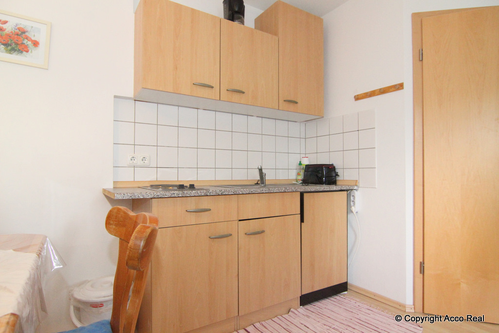 Küchenzeile mit Holzfronten, Spüle und gefliestem Wandbereich; davor steht ein Holzstuhl.