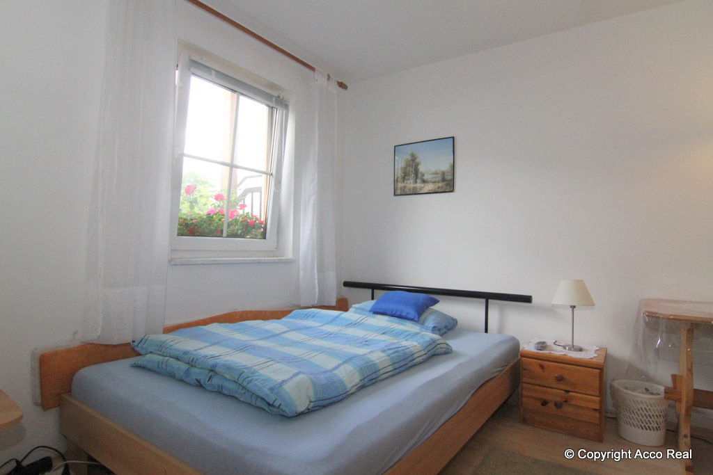 Bett mit blau gemusterter Bettwäsche neben einem Fenster und einem kleinen Holznachttisch.