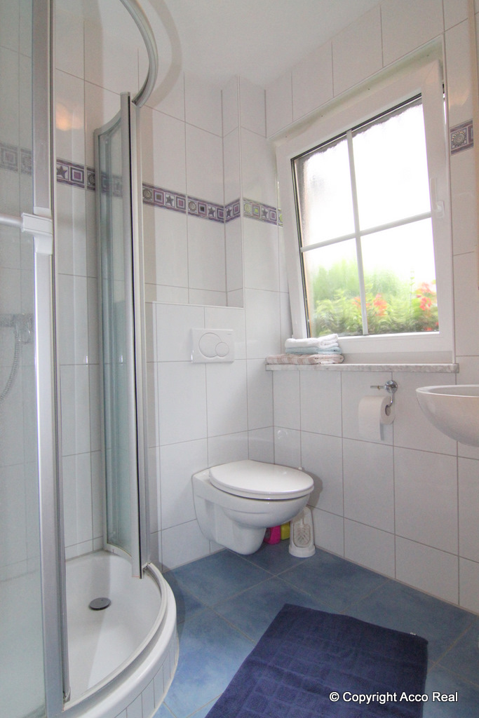 WC unter dem Badezimmerfenster, daneben eine verglaste Duschkabine und ein Waschbecken.