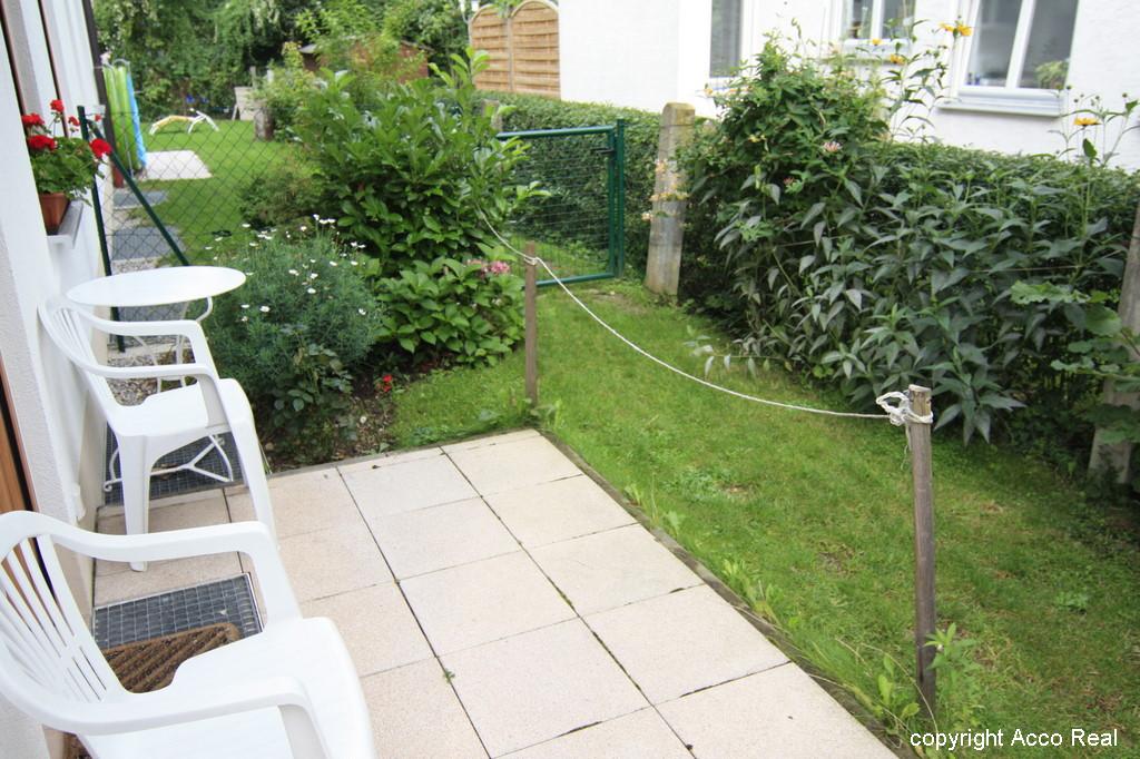 Terrassenfläche mit weißen Stühlen neben einer Rasenfläche mit Sträuchern.